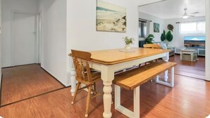A dining area with a wooden table and chairs, a bench, a painting on the wall, and a view of a mountain through a window. - BROOMS HEAD PET FRIENDLY BEACH PAD with authentic retro arcade games machine. 🐶 (Brooms Head)