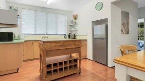A kitchen with wooden cabinets, a stainless steel refrigerator, a wooden table, and a window with blinds. - BROOMS HEAD PET FRIENDLY BEACH PAD with authentic retro arcade games machine. 🐶 (Brooms Head)