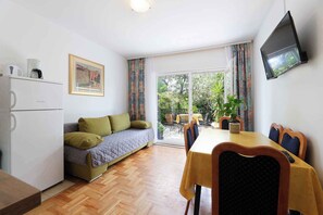A compact living space with a kitchenette, a sofa, a dining table with chairs, a TV mounted on the wall, and a sliding glass door leading to an outdoor area. - Holiday apartment with air conditioning and garden (Jadranovo)