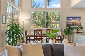 Ein Wohnzimmer mit Esstisch, Stühlen, einer Couch und großen Fenstern mit Blick auf Bäume und einen Sonnenuntergang.
