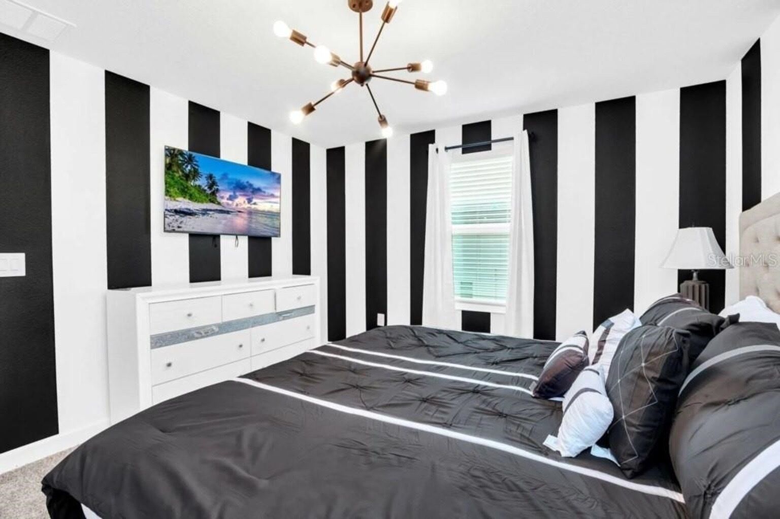 A bedroom with a black and white striped wall design, a bed with a black and white striped bedspread, a white dresser, a television on the wall, and a window with blinds.