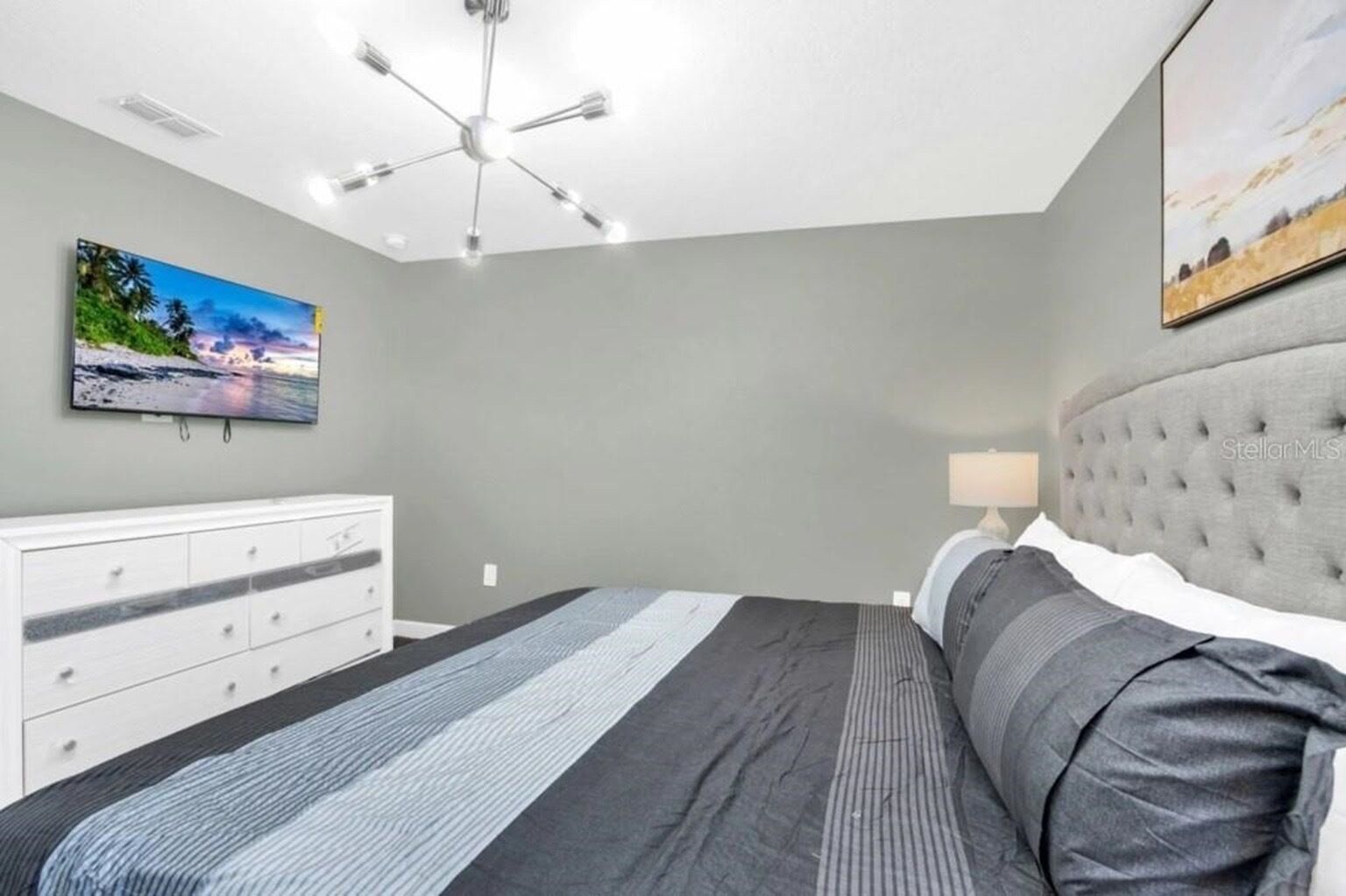 A bedroom with a bed, a dresser, a ceiling fan, a painting on the wall, and a lamp on the nightstand.