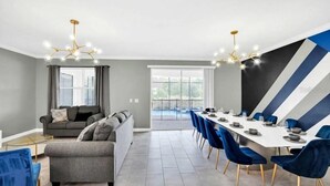 A modern living room with a sofa, a dining area with a long table and chairs, and a large window. - Beautiful 9-bedroom house with AC in serene Kissimmee , Bring the whole Family ! (Kissimmee)