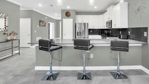 A modern kitchen with white cabinets, a stainless steel refrigerator, and a microwave. There are three bar stools with black seats and chrome legs. - Beautiful 9-bedroom house with AC in serene Kissimmee , Bring the whole Family ! (Kissimmee)