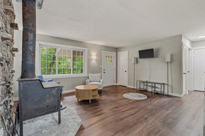A spacious living room with a stone wall, a fireplace, a round coffee table, a white armchair, and a large window with a view of greenery.