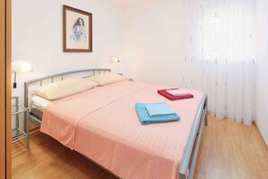 A neatly made bed with a pink bedspread, a blue folded towel, and a red towel on the side. There is a wall-mounted picture and a bedside lamp. - Holiday flat pleasantly overlooking the courtyard and garden (Jadranovo)
