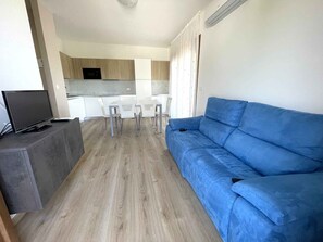 Ein Wohnzimmer mit blauem Sofa, einem Fernseher auf einem Schrank, einem Esstisch mit Stühlen und einer Küche im Hintergrund.