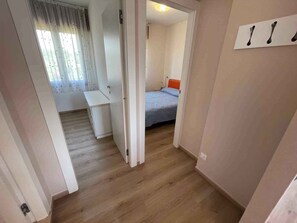 Een smalle gang die leidt naar een slaapkamer met een bed, een bureau en een stoel.