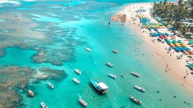 An aerial view of a beach with boats, clear turquoise water, and a row of beach umbrellas. - Sunny Hall By Flat na Praia (Ipojuca)