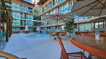 Zona de piscina de hotel con varios niveles de balcones, mobiliario exterior y sombrillas.