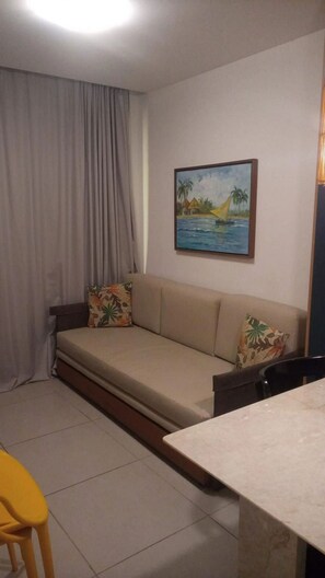 Una habitación con un sofá, un cuadro de una escena tropical, una cortina y una silla amarilla.