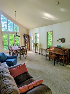 A spacious living room with a piano, a dining area, and a large window with a view of greenery. - Lovely cottage close to vineyards and beaches. (Benton Harbor)