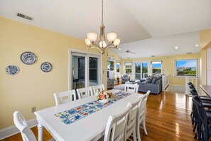 A dining area with a white table, chairs, and a chandelier. There are decorative plates on the wall and a view of the outside through large windows. - Northshore Lookout: Golf & Beach Retreat (Nags Head)