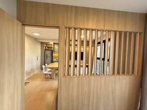 Un interior moderno con un separador de madera, una zona de comedor con sillas blancas y una cocina con refrigerador y armarios.