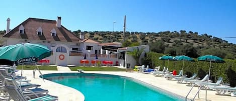 Una piscina con tumbonas y sombrillas, un edificio de techo con tejas y un paisaje montañoso al fondo.