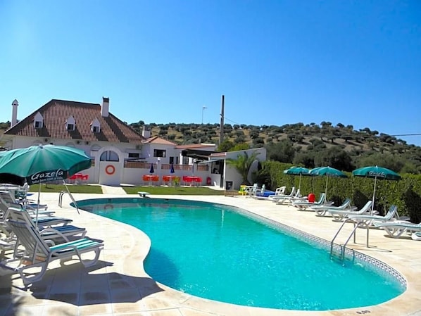 A swimming pool with lounge chairs and umbrellas, a building with a tiled roof, and a hilly landscape in the background. - Jardim Hotel (Santo Tirso)