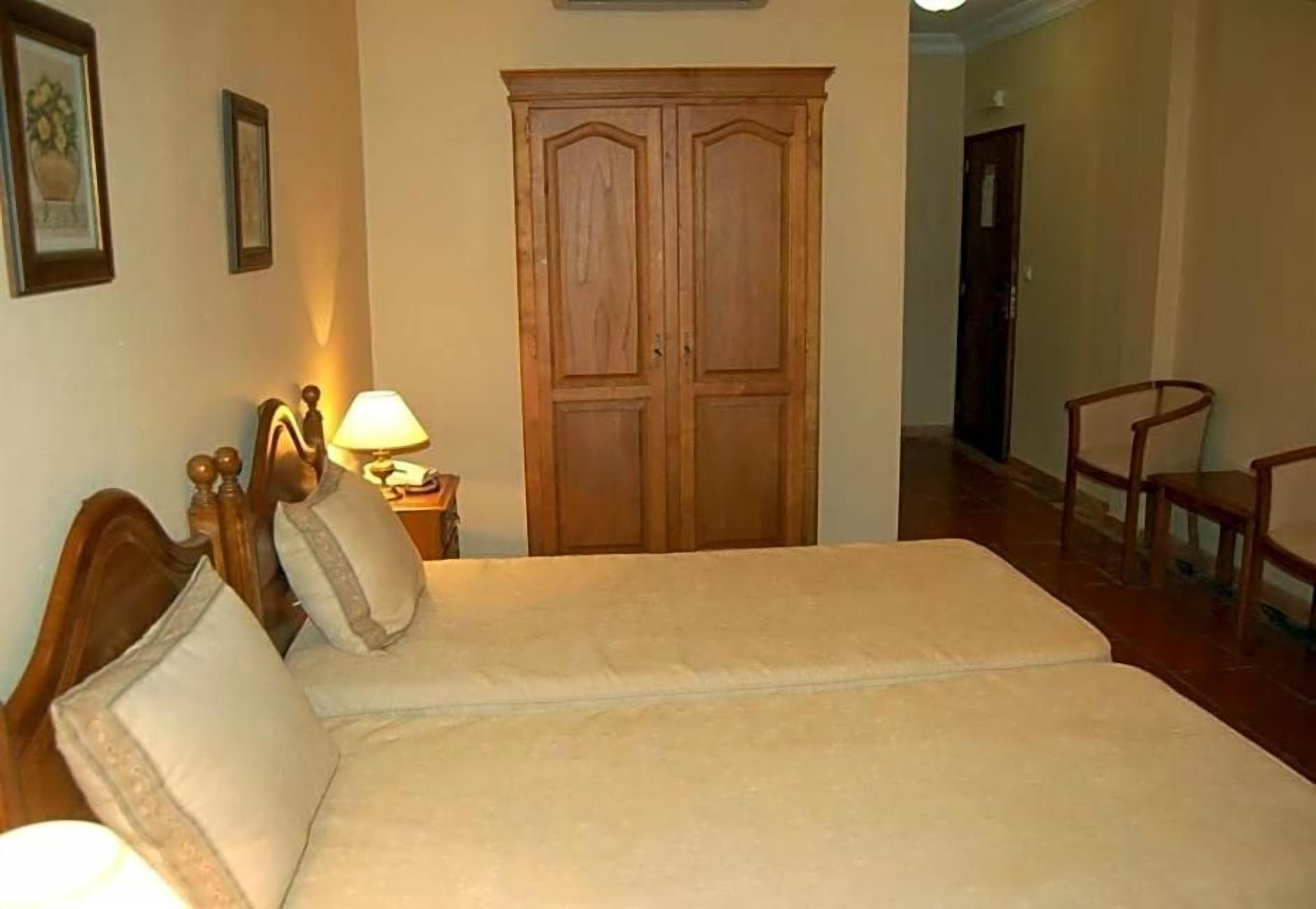 A bedroom with a bed, a wooden wardrobe, a nightstand with a lamp, and a chair.
