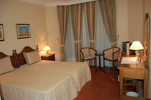 A hotel room with two beds, a desk with a lamp, a chair, and a television. - Jardim Hotel (Santo Tirso)