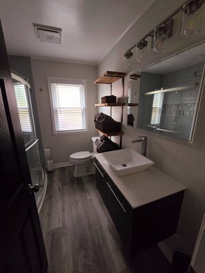 A bathroom with a white sink, dark cabinets, a mirror, and a window with blinds. - Fully Furnished Insurance Housing 3BR Home (Detroit)