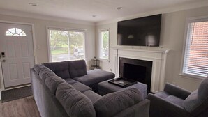 En stue med en hjørnesofa, pejs, tv ophængt på væggen og en dør med et vindue.