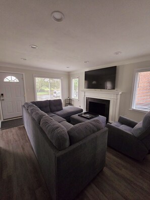 A living room with a sectional sofa, a fireplace, a TV mounted on the wall, and a door with a window. - Fully Furnished Insurance Housing 3BR Home (Detroit)
