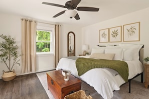 Ein Schlafzimmer mit Bett, Deckenventilator, Fenster mit Vorhängen, Nachttisch und Spiegel. An den Wänden hängen gerahmte Pflanzenbilder.