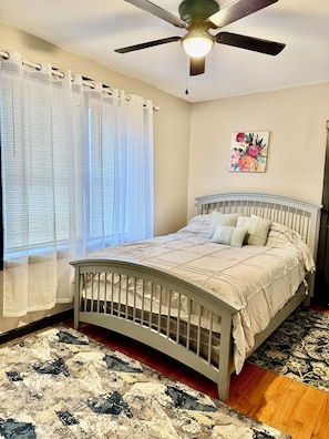 Ein Schlafzimmer mit Bett, Deckenventilator, Fenster mit durchscheinenden Vorhängen, einem floralen Gemälde an der Wand und einem gemusterten Teppich.