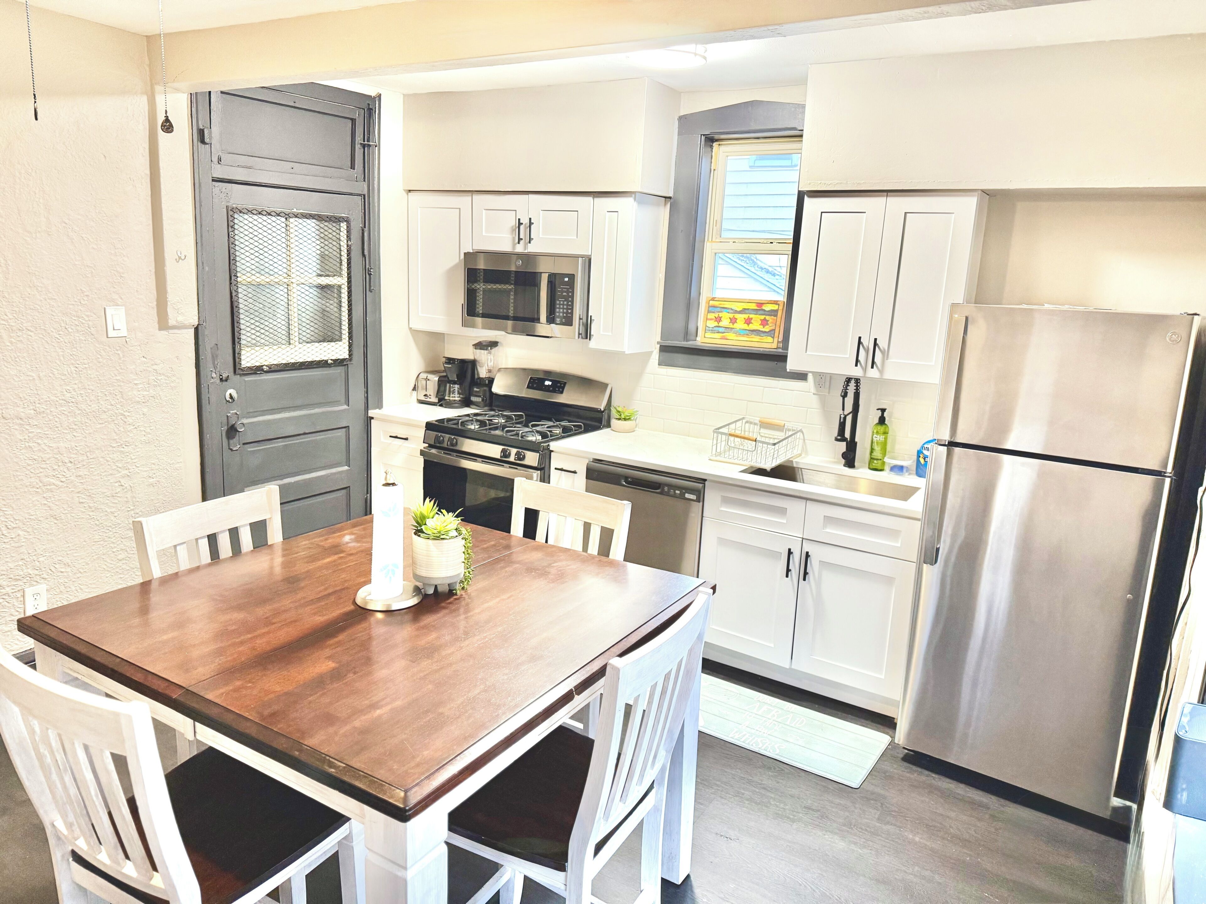 A compact kitchen with white cabinets, a stainless steel refrigerator, a microwave, and a stove. There is a dining table with chairs and a small plant on it.