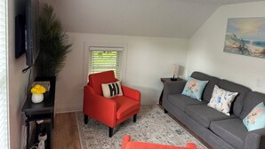 A living room with a red armchair, grey sofa, a flat-screen TV, a potted plant, a painting, and a window with blinds. - MedCity Suite—2BR/1BA Near Mayo Clinic (Rochester)