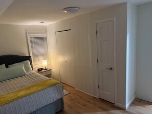 Un dormitorio con cama, mesita de noche con lámpara, ventana con persianas y una puerta.