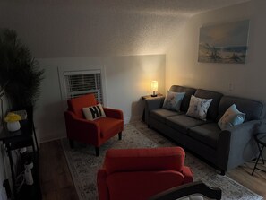 A living room with a red armchair, a grey sofa, a small table with a lamp, and a painting on the wall. - MedCity Suite—2BR/1BA Near Mayo Clinic (Rochester)