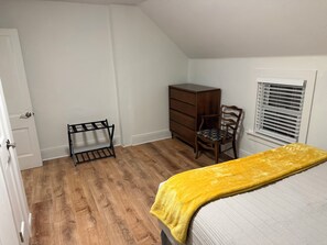 Ein Schlafzimmer mit Bett, Kommode, Stuhl, Fenster mit Jalousien und einer Tür.
