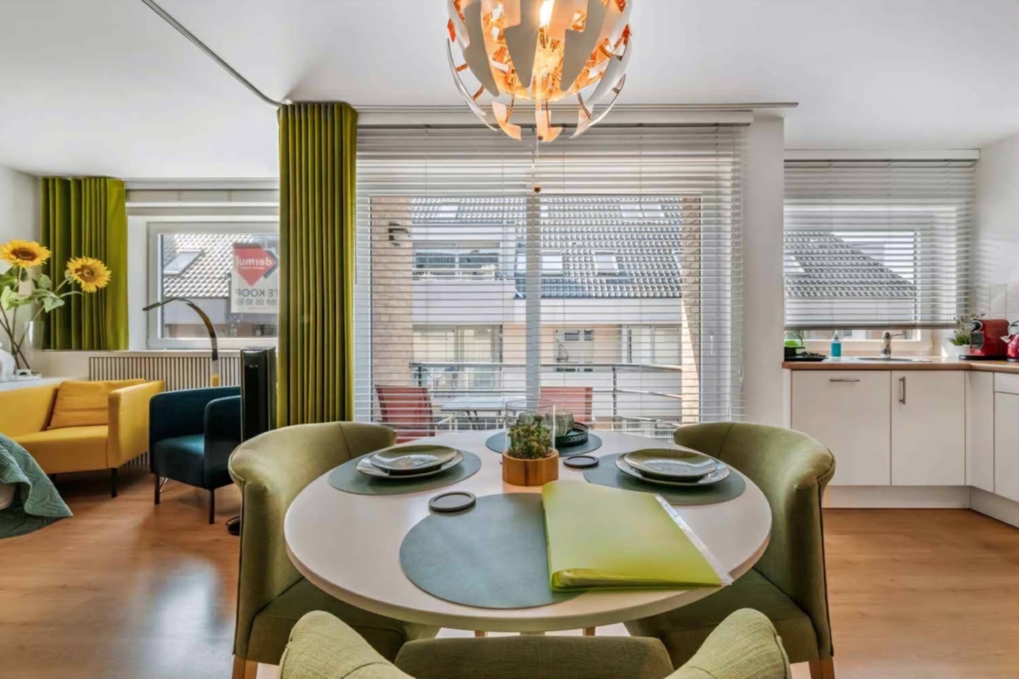 A modern living room with a dining table, green curtains, a yellow sofa, and a view of the city through large windows.