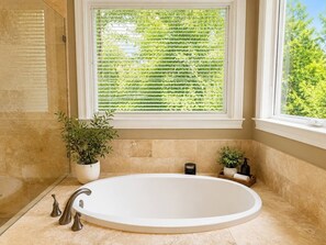 A bathroom with a large oval bathtub, a potted plant, a window with blinds, and a towel rack. - Hill Country Oasis with Pool, Spa & Views (Austin)