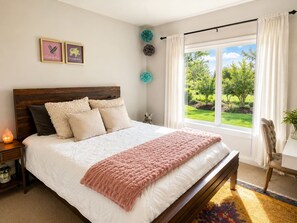 A bedroom with a bed, a nightstand, a window with curtains, and a view of greenery outside. - Hill Country Oasis with Pool, Spa & Views (Austin)