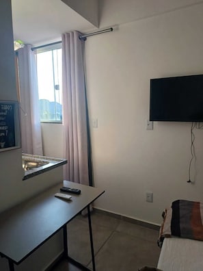 Habitación de hotel con cama, televisión montada en la pared, ventana con cortinas, escritorio con control remoto y un lavamanos pequeño.