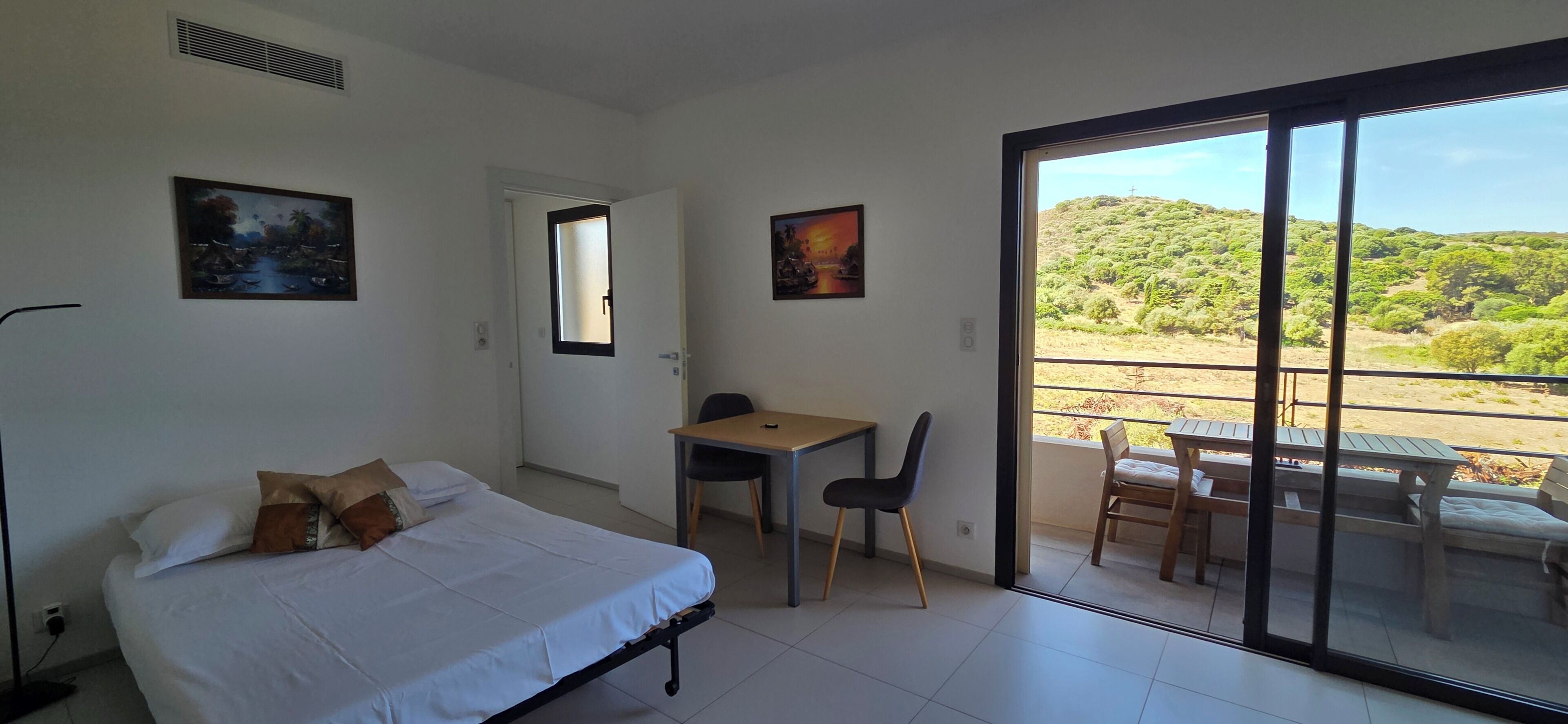 Une chambre avec un lit, un bureau et une chaise, un balcon avec une table et des chaises, et deux tableaux accrochés aux murs.