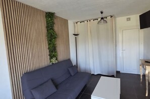 A modern living room with a dark gray sofa, a white coffee table, a floor lamp, and a wall with vertical wooden slats and a green plant. - Dolce & Vita Studio D3 with ocean and pool views (Propriano)