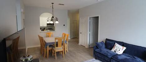 A living room with a blue sofa, a dining table with chairs, a ceiling fan, and a kitchen area in the background.