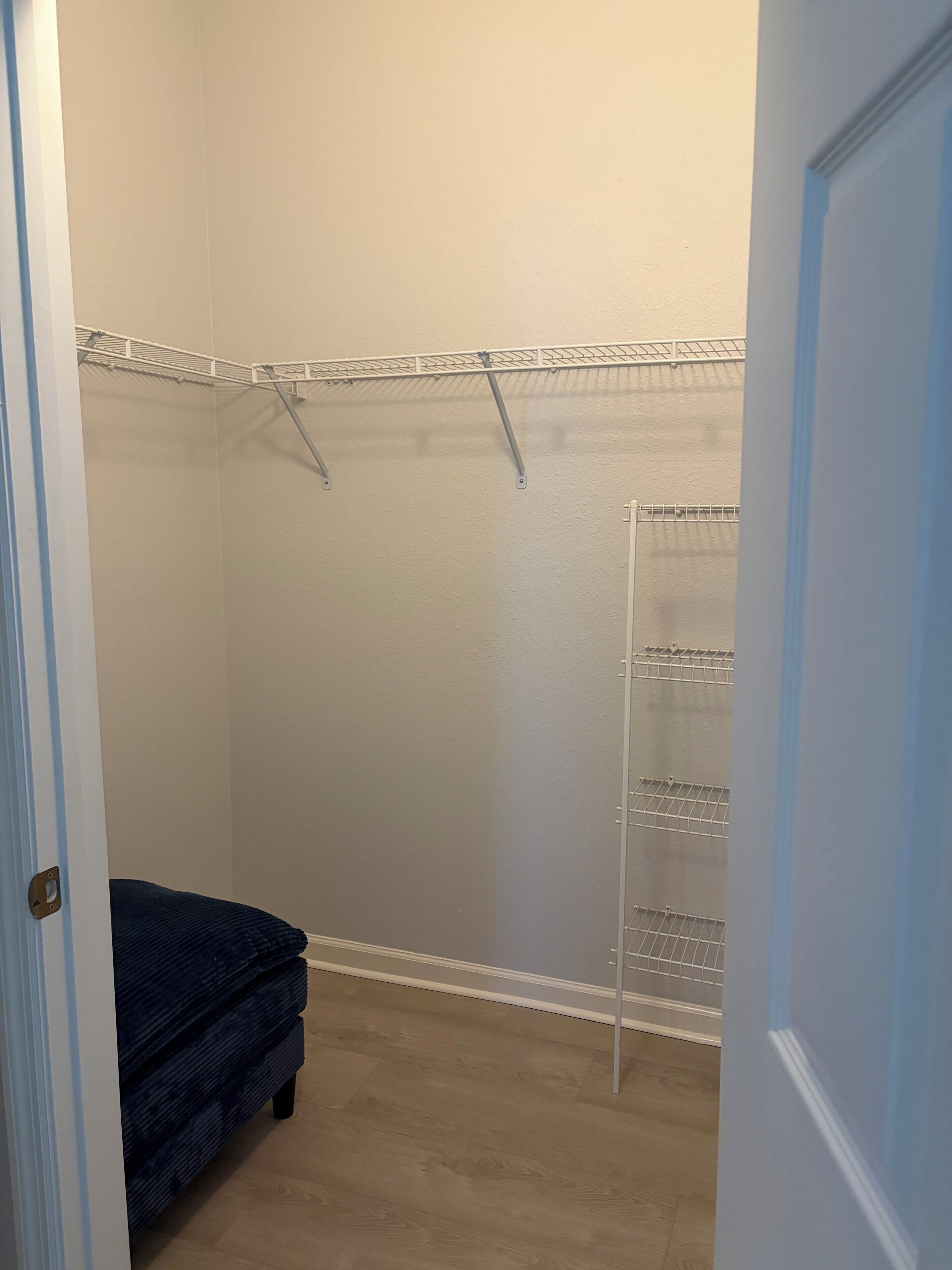 A walk-in closet with a metal shelving unit, a dark-colored bench, and a plain wall.