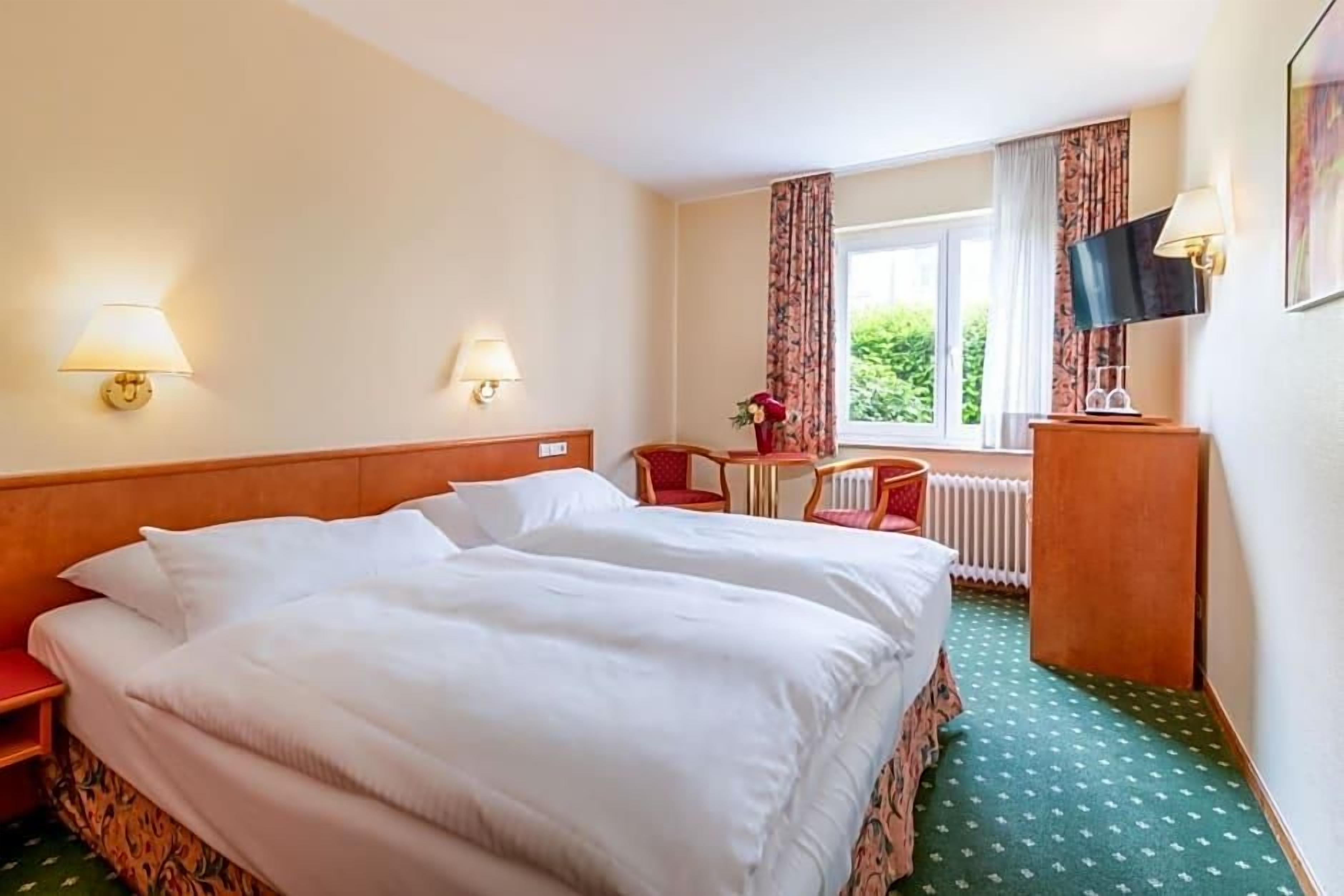 Ein Hotelzimmer mit Doppelbett, zwei Nachttischlampen, einem kleinen Tisch mit Stuhl, einem Fernseher und einem Fenster mit Vorhängen.