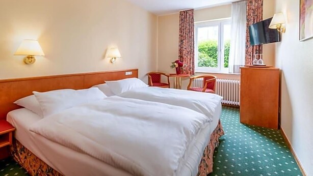 Et hotelværelse med dobbeltseng, to sengeborde med lamper, et lille bord med en stol, et fjernsyn og et vindue med gardiner.