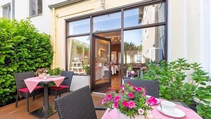 En udendørs terrasse med fletstole og et spisebord, et stort vindue med udsigt ind til bygningen, og krukkeplanter.