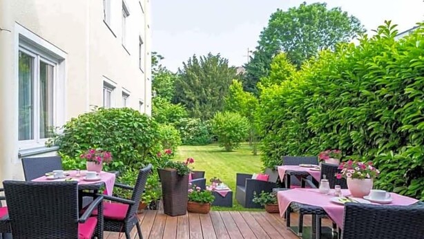 An outdoor patio with wicker furniture and pink tablecloths, surrounded by lush greenery and a view of a lawn. - Alte Post (Krefeld)