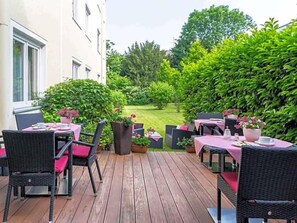 An outdoor patio with wicker furniture and pink tablecloths, surrounded by lush greenery and a view of a lawn. - Alte Post (Krefeld)