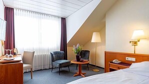 Quarto de hotel com cama, escrivaninha com luminária, cadeira, pequena mesa com planta e janela com cortinas.