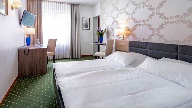 Quarto de hotel com uma cama grande, uma escrivaninha com computador, uma cadeira e uma pequena mesa com um vaso de flores.