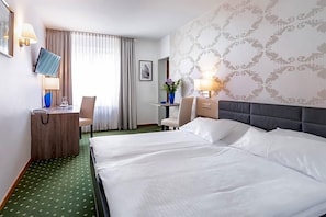 Ett hotellrum med en stor säng, ett skrivbord med en dator, en stol och ett litet bord med en vas med blommor.