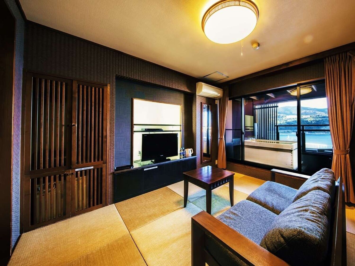 A room with tatami flooring, a sofa, a television, and a view of a body of water through large windows.