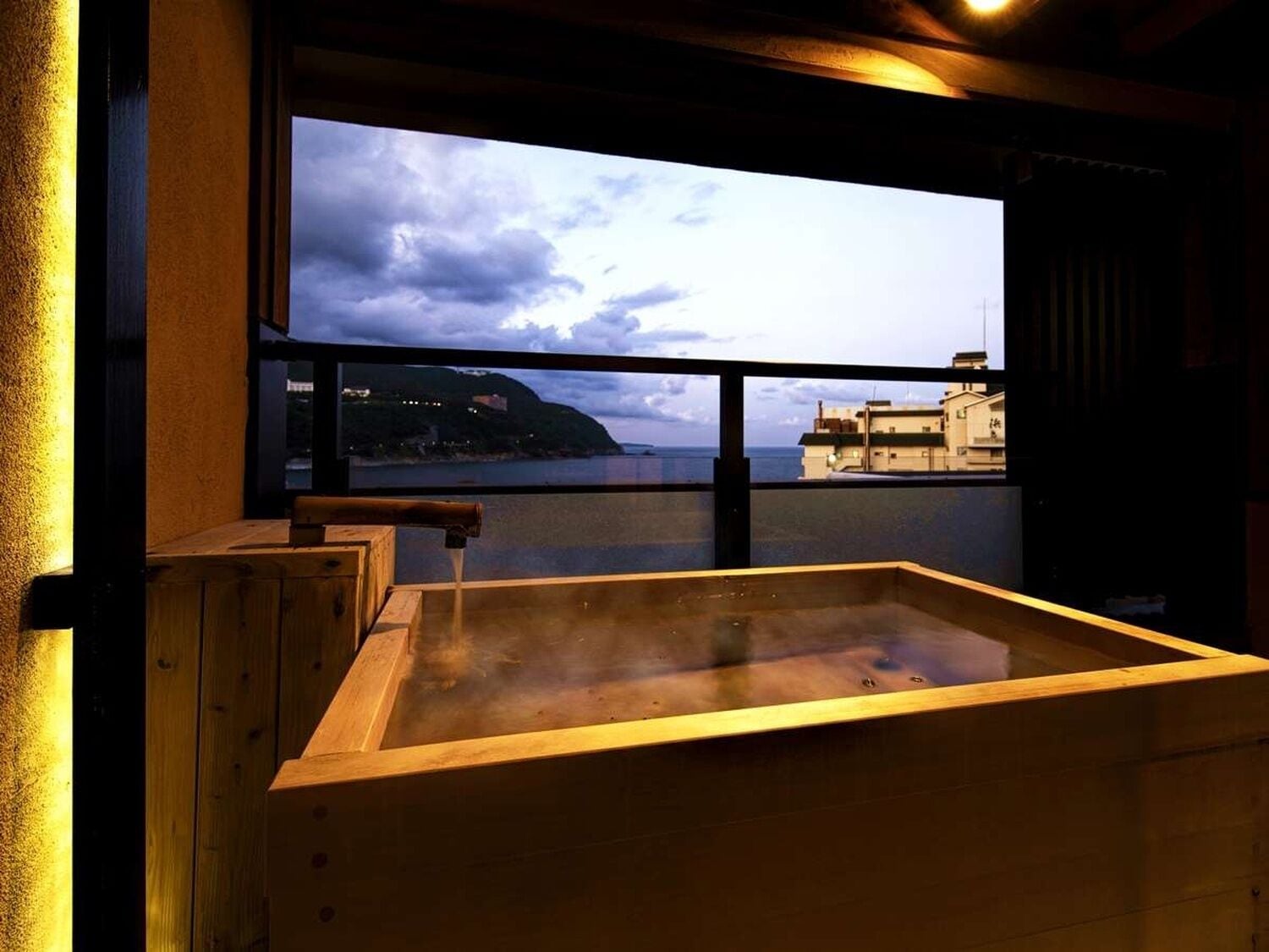 A room with a wooden sauna, a large window overlooking a coastal view, and a view of buildings across the water.